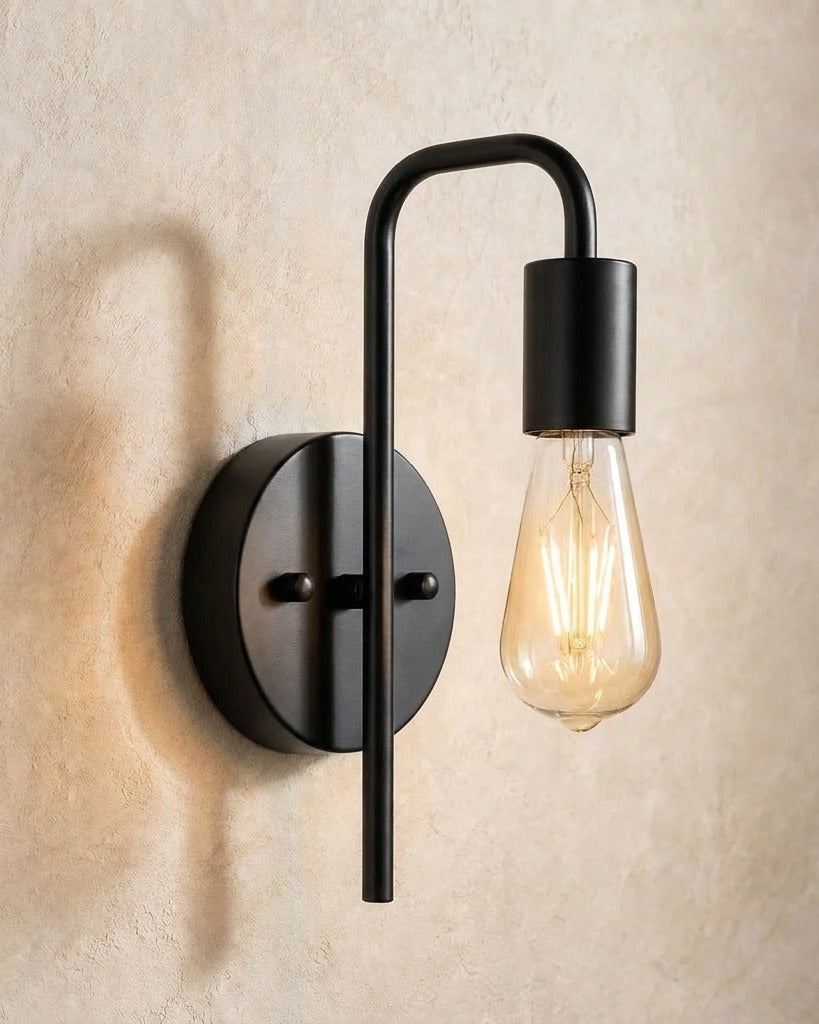 The Filament black industrial wall sconce with exposed bulb, lit, mounted on a lime-washed plaster wall — Selden Home product photography