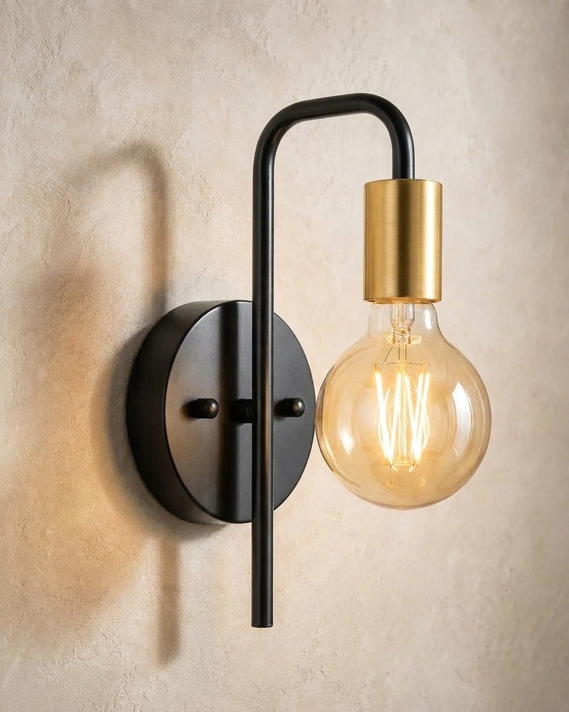The Filament black and gold industrial wall sconce with exposed bulb, lit, mounted on a lime-washed plaster wall — Selden Home product photography