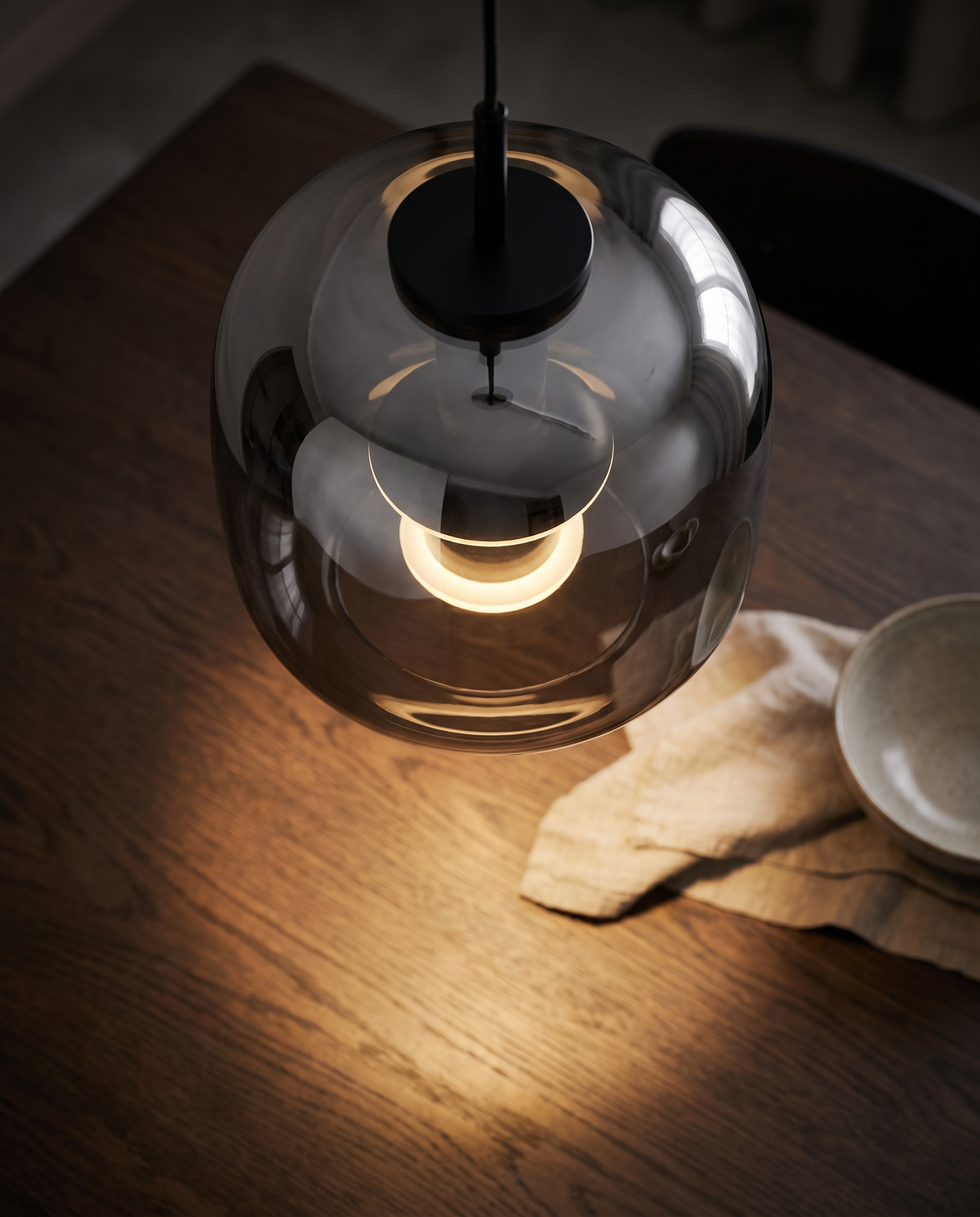 The smoked grey Capsule Pendant seen from above, its light pooling on an oak table
