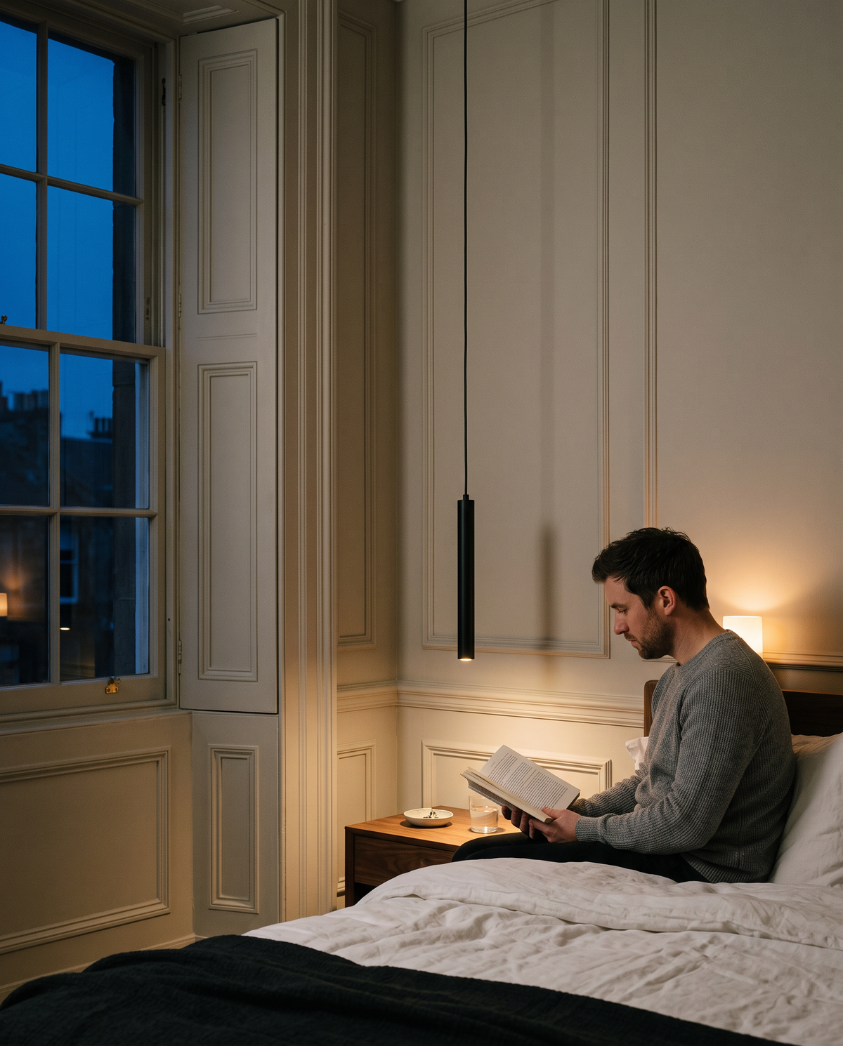A man reading on the edge of the bed under a Plumb pendant